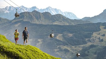 Zwei Personen wandern am Berg, im Hintergrund sieht man die Gondeln hinauffahren | © saalbach.com, Karin Pasterer